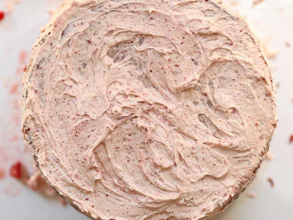 A close-up of a vegan strawberry cake topped with creamy, swirled frosting. The frosting has a light pink hue with small red specks, giving it a textured appearance. The cakes round shape is centered in the image.